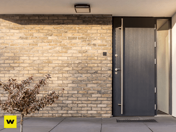 Moderne Haustür aus Aluminium vor heller Klinkerfassade mit Pflanze und Außenbeleuchtung.